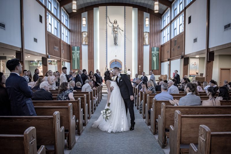 A bride and groom kiss at an altar in a modern church as guests smile and clap, with natural light streaming through windows.