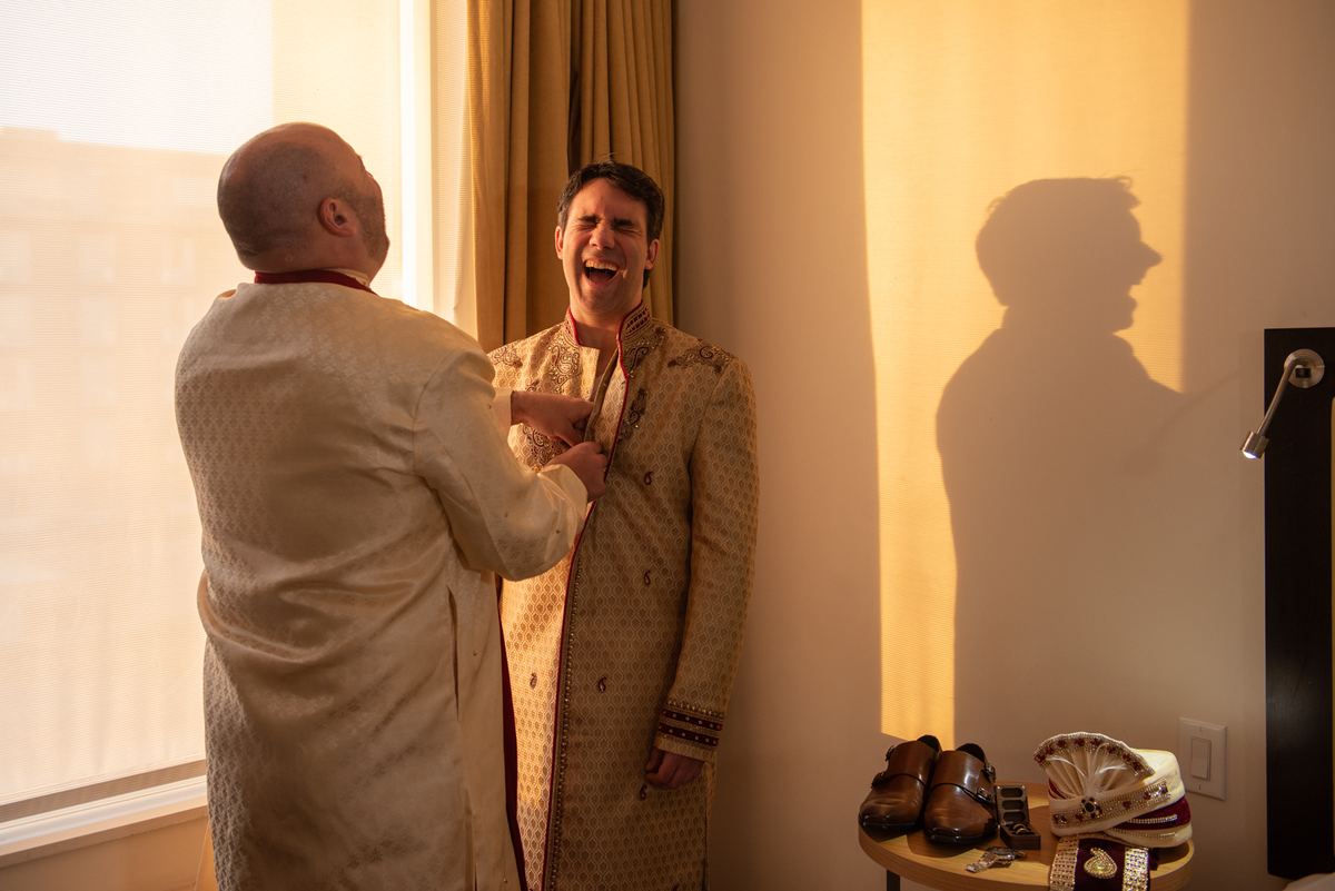 A groom laughs as a family member adjusts his sherwani near a window with ceremonial shoes and turban on a table.