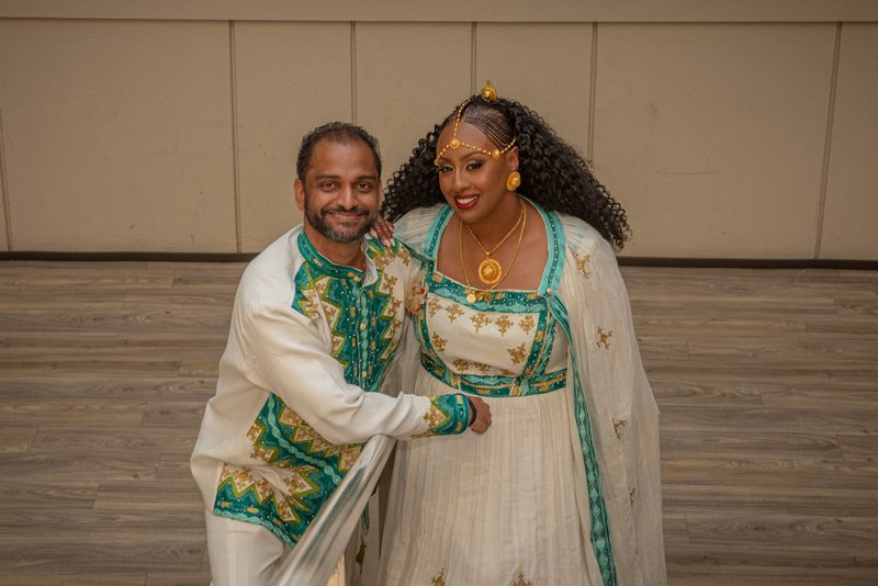 Salem and Shannon, dressed in ornate traditional attire, pose closely together with smiles in a warmly lit indoor space.