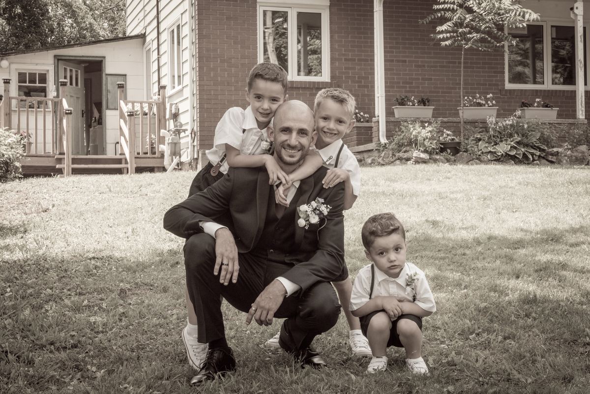 A groom crouches in a grassy yard with three young boys dressed formally, embracing him warmly as the youngest sits nearby.
