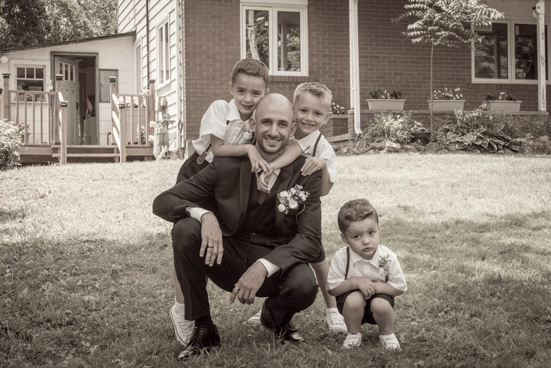 A groom crouches in a grassy yard with three young boys dressed formally, embracing him warmly as the youngest sits nearby.