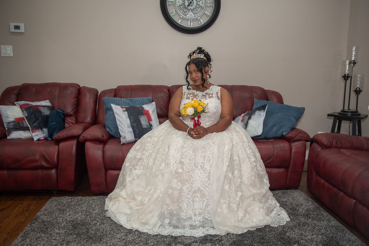 A woman in a white lace gown and tiara sits on a red leather sofa holding yellow and white flowers, near a wall clock.