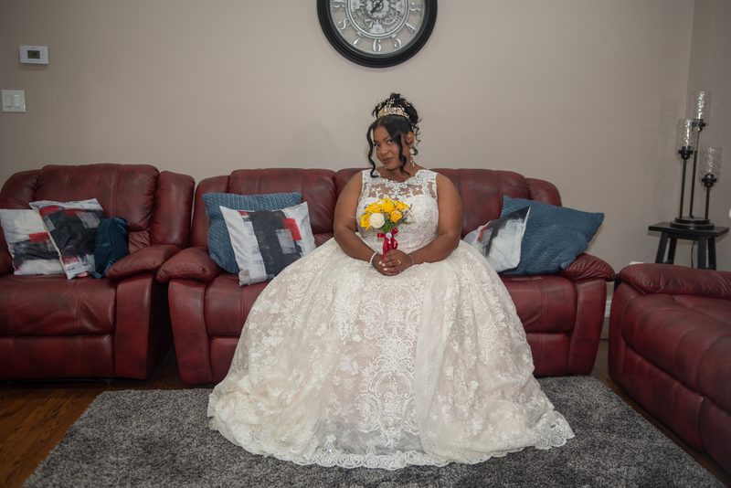 A woman in a white lace gown and tiara sits on a red leather sofa holding yellow and white flowers, near a wall clock.