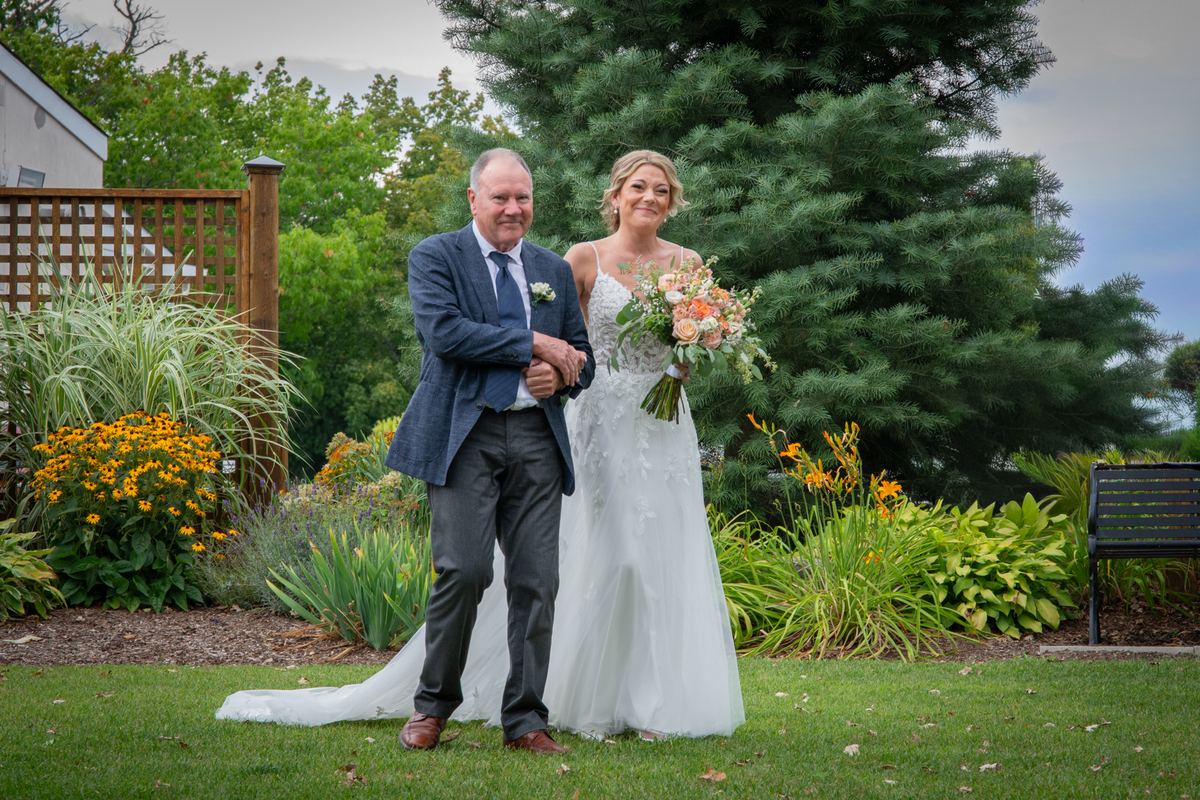 A bride in a white gown and floral bouquet stands with her smiling father, hand in hand, before the garden ceremony.
