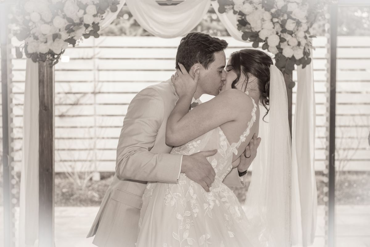 A bride and groom kiss tenderly beneath an arbor adorned with flowers and fabric, bathed in soft monochromatic light.
