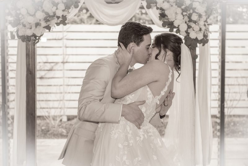 A bride and groom kiss tenderly beneath an arbor adorned with flowers and fabric, bathed in soft monochromatic light.