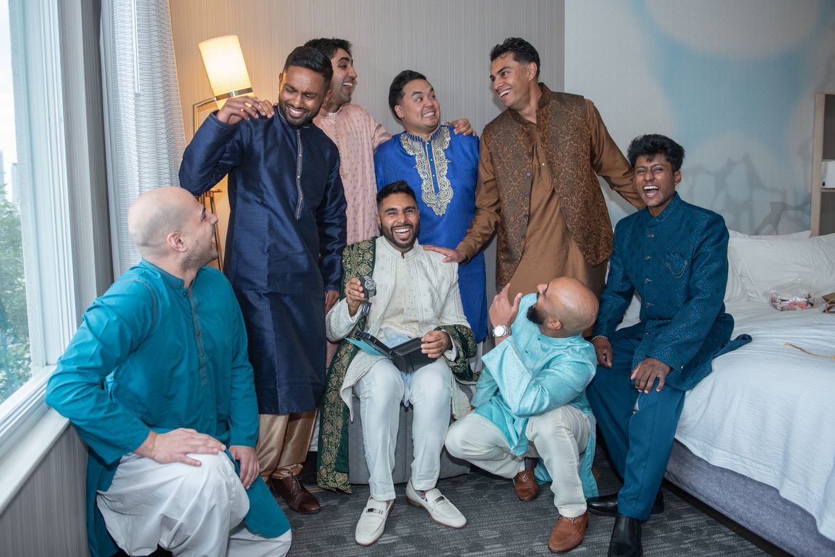 South Asian men in traditional attire gather and laugh in a hotel room, illuminated by natural and warm indoor light.
