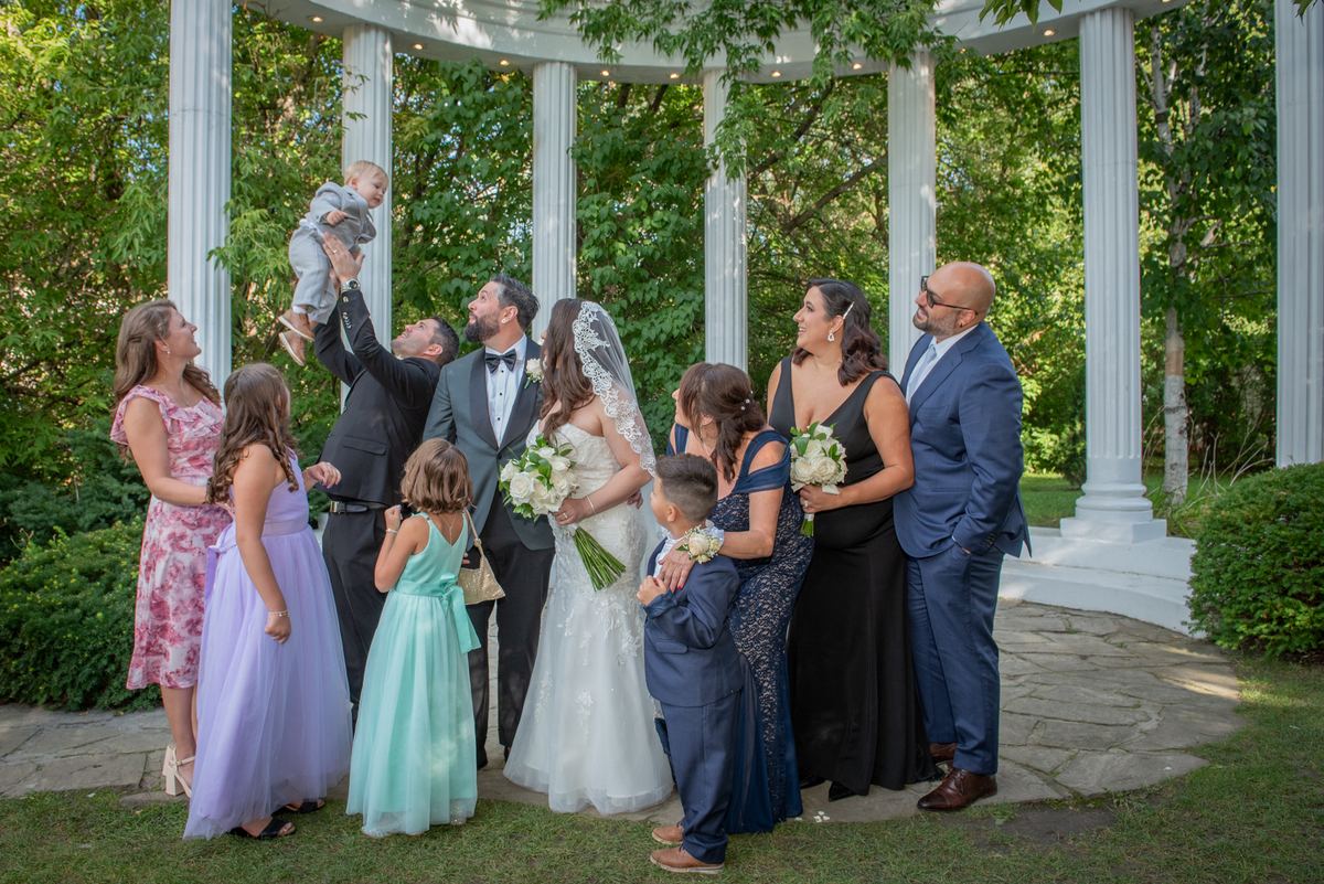 Bride and groom surrounded by smiling family, with a man lifting a baby above their heads joyfully near a white gazebo.