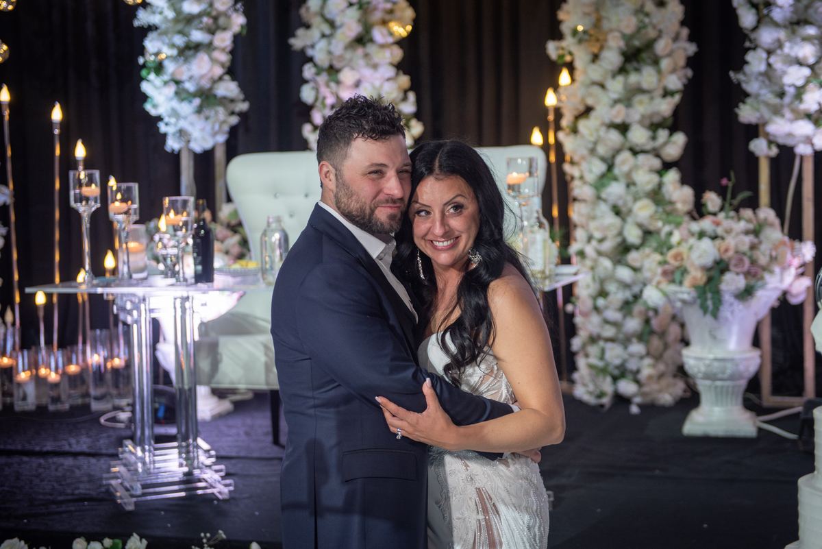A newlywed couple embraces warmly in a candle-lit, elegantly decorated space surrounded by flowers and table settings.
