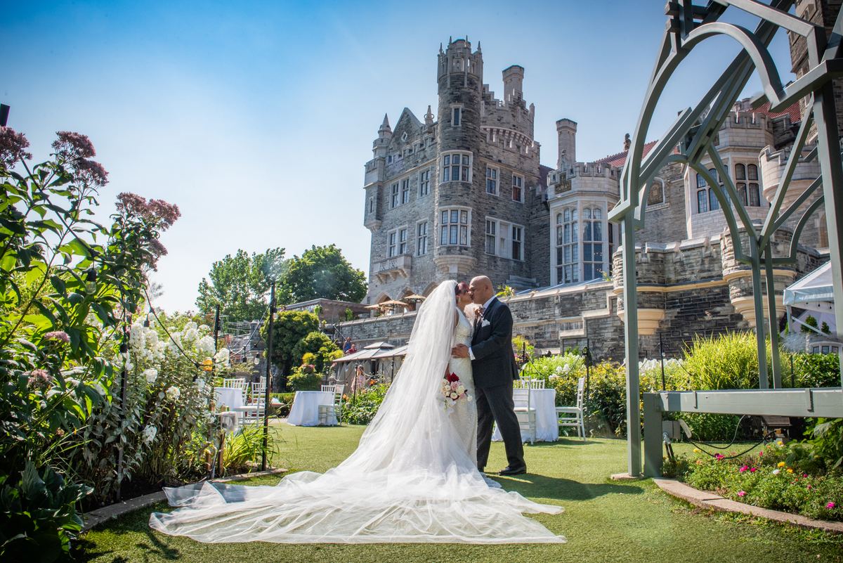 A bride and groom kiss on a sunlit lawn near a floral arch, surrounded by stone towers and garden foliage.