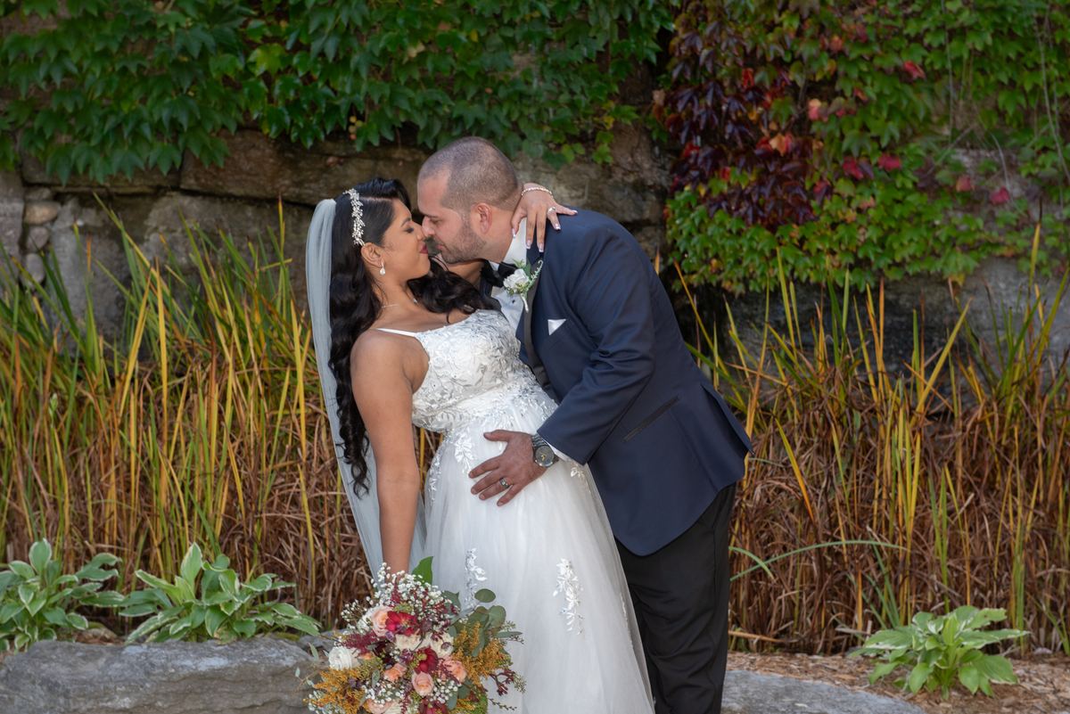 A bride and groom kiss tenderly outdoors, surrounded by lush greenery and autumn leaves, bathed in soft natural light.
