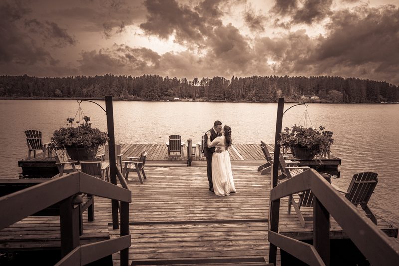 A newlywed couple embraces on a wooden dock by a calm lake at sunset, surrounded by hanging planters and empty chairs.