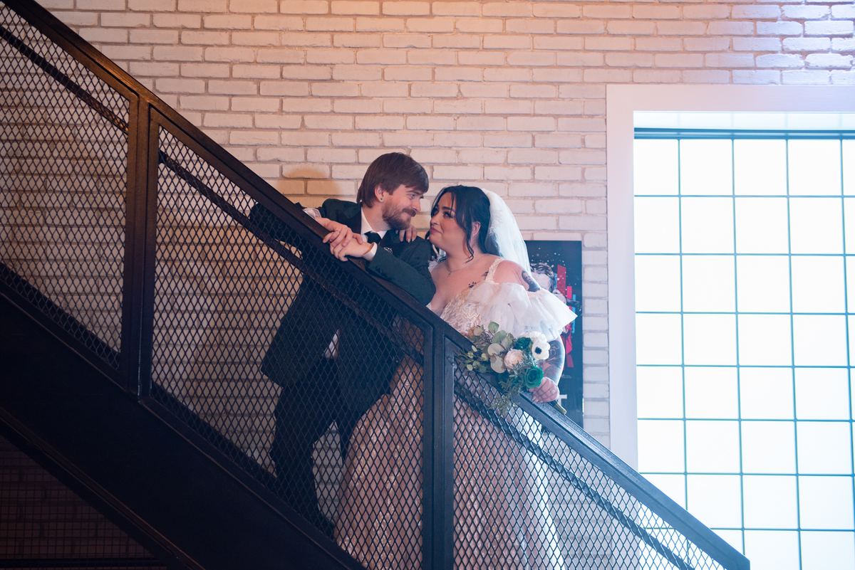 Bride and groom lean against a metal railing, gazing at each other warmly in soft light from a large window.