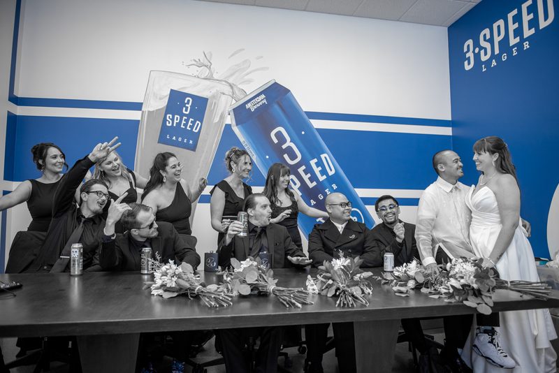 A smiling bride and groom stand beside a table with bouquets and beer cans as guests toast in celebration.