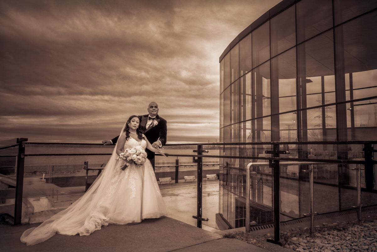 A bride in a flowing gown and groom in a tuxedo pose on a modern rooftop terrace at sunset, surrounded by glass architecture.