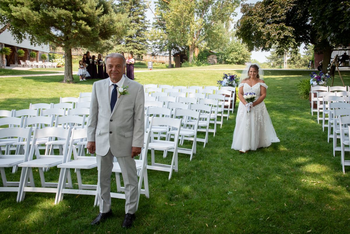 A bride in a white gown walks down a grassy aisle toward the camera, flanked by chairs and guests under trees.