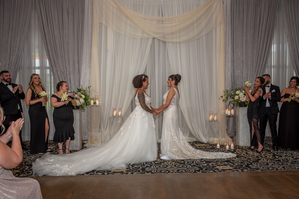 Two brides in white gowns hold hands and smile as guests applaud during an intimate ceremony with candlelit pedestals.