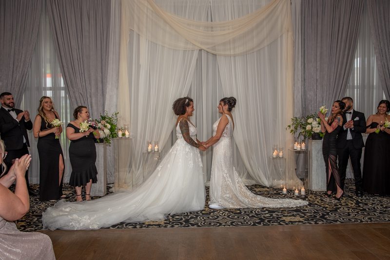 Two brides in white gowns hold hands and smile as guests applaud during an intimate ceremony with candlelit pedestals.