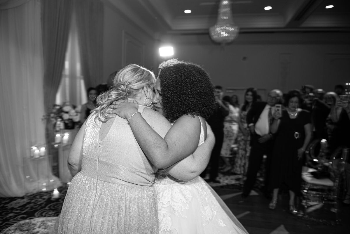 A bride and family member embrace warmly at a wedding reception, surrounded by guests and illuminated by ambient lights.