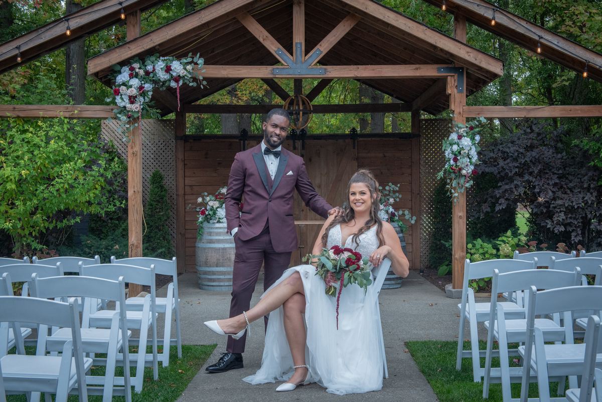 A smiling groom stands beside his seated bride under a floral-adorned wooden arbor at an outdoor wedding.