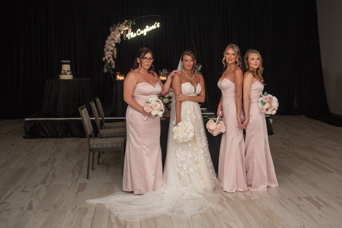 A bride and her bridesmaids pose with floral bouquets in a warmly lit reception space, behind them a cake and a glowing sign.