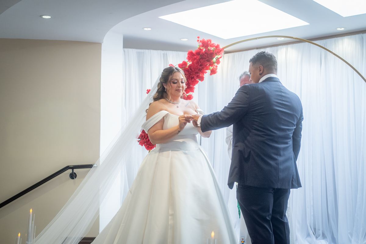 A bride and groom exchange rings under an arched floral decoration, surrounded by minimalist decor and soft natural light.