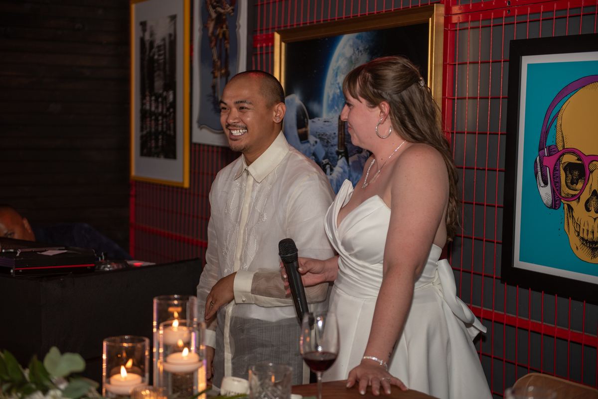 A smiling groom in a barong tagalog and his bride in a white gown hold microphones at a candlelit table with modern art.