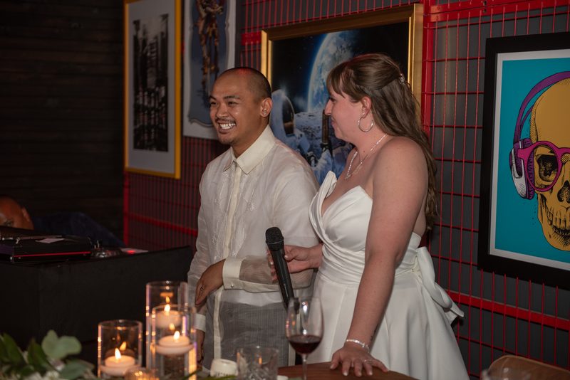 A smiling groom in a barong tagalog and his bride in a white gown hold microphones at a candlelit table with modern art.