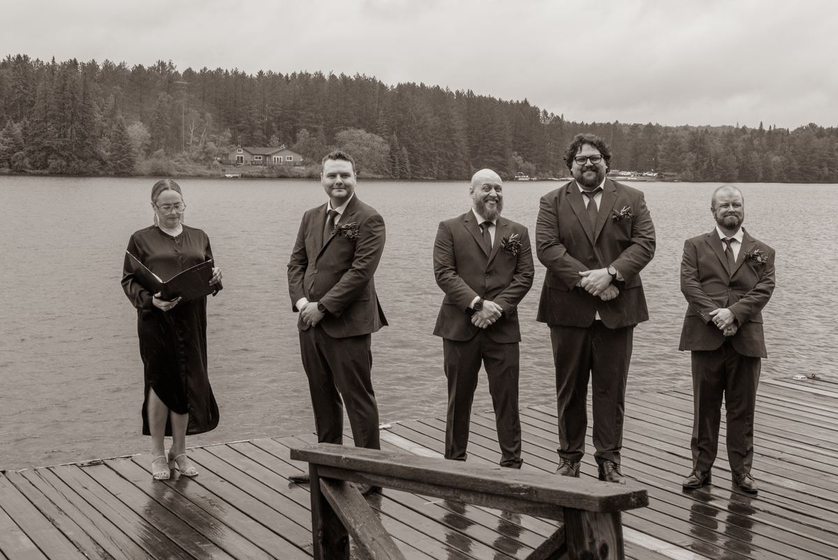 A wedding ceremony takes place on a wet dock by a lake under an overcast sky, with the officiant and groomsmen in suits.