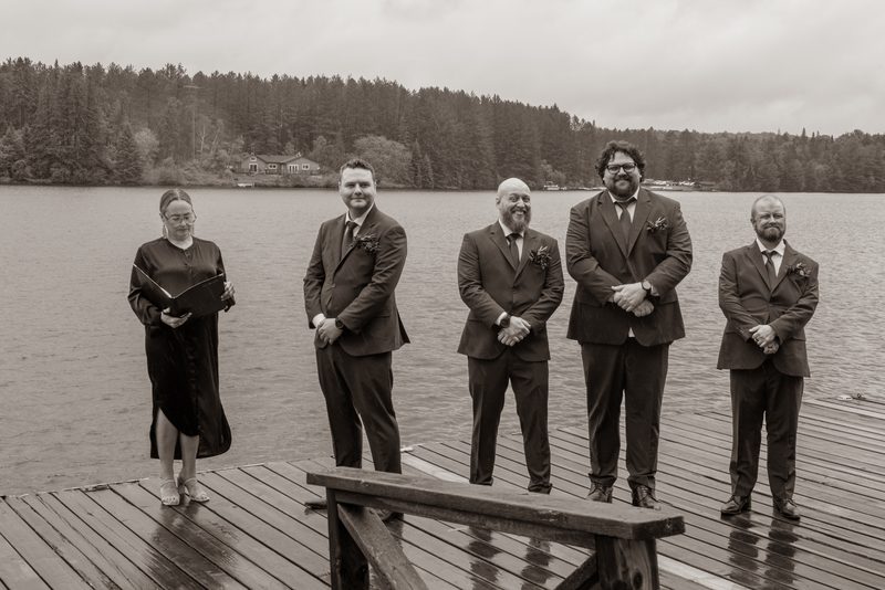 A wedding ceremony takes place on a wet dock by a lake under an overcast sky, with the officiant and groomsmen in suits.