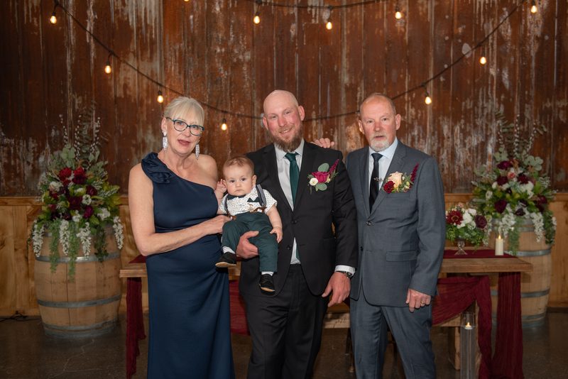 A groom holds his baby as parents look on at a wedding celebration with warm lights and formal attire.