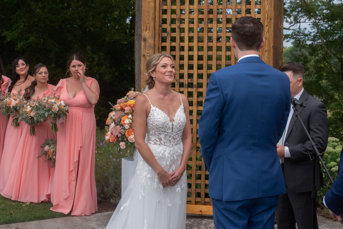 Bride in white lace gown looks at groom in blue suit during outdoor ceremony with floral arrangements and bridesmaids.