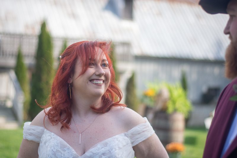 A smiling red-haired bride in a white off-the-shoulder gown looks at her groom near a rustic barn surrounded by greenery.