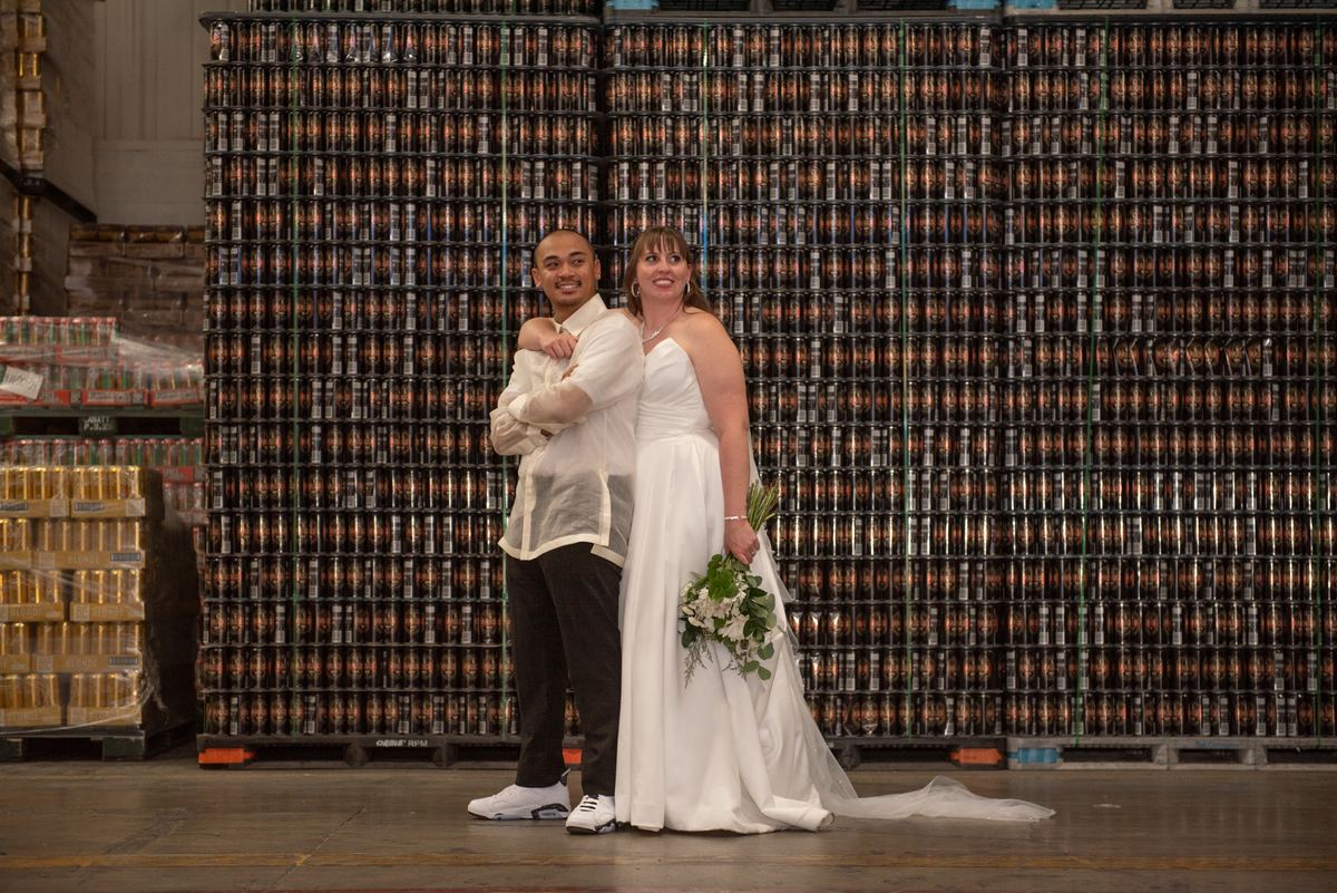 A newlywed couple in formal attire stands before a wall of stacked beer cans in a warehouse, smiling at the camera.