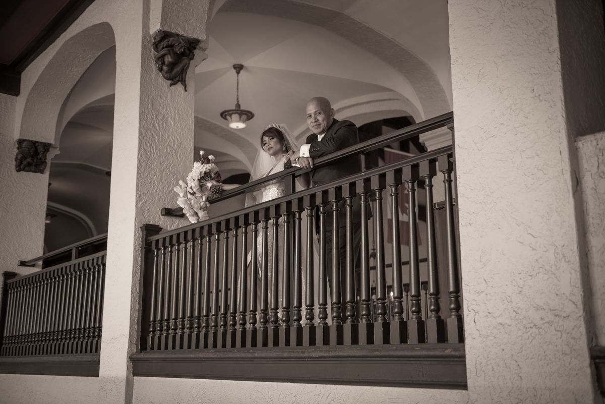 A bride and groom stand on an ornate balcony, gazing at guests below, framed by arched architecture.