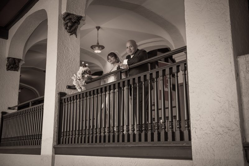 A bride and groom stand on an ornate balcony, gazing at guests below, framed by arched architecture.