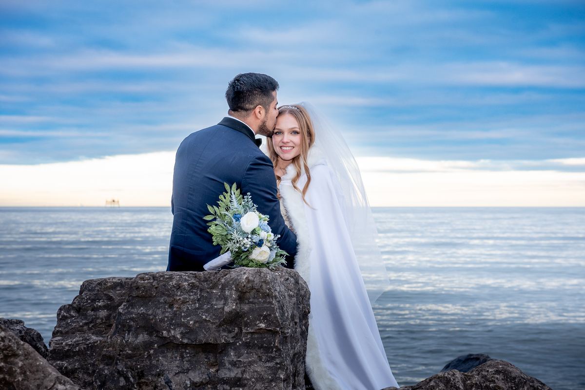 A newlywed couple stands on rocky cliffs, the groom kissing the bride's temple as she smiles at the camera.