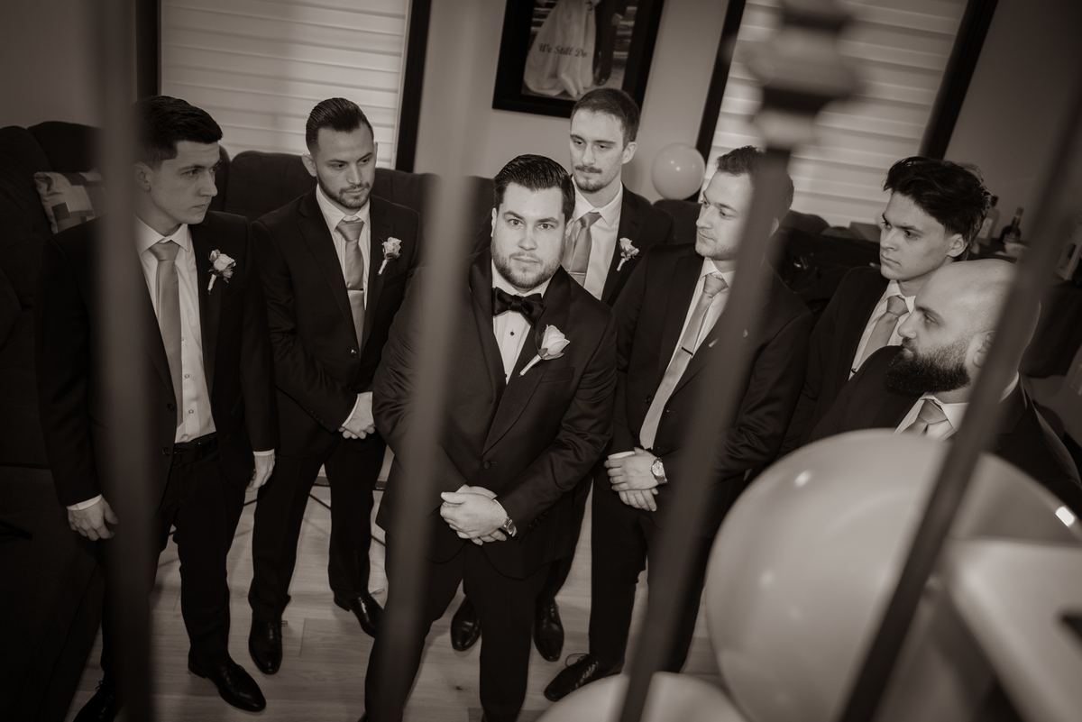 A group of groomsmen in formal suits stands around the groom, who looks directly forward with a serious expression.