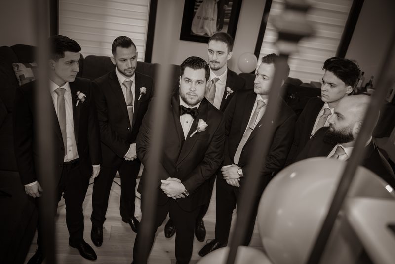 A group of groomsmen in formal suits stands around the groom, who looks directly forward with a serious expression.