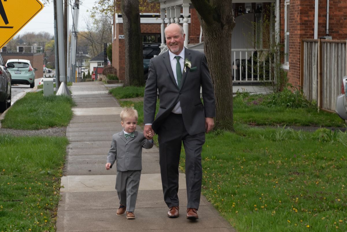 An older man and a child in matching gray suits walk hand-in-hand down a suburban sidewalk, smiling.