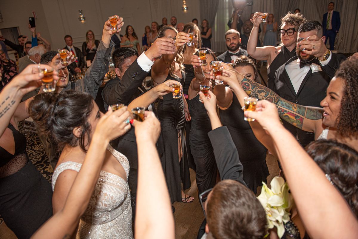 Guests in formal attire raise shot glasses in unison during a joyful wedding toast, their faces illuminated by warm lighting.
