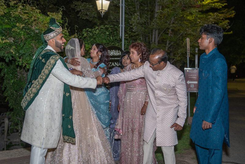 A groom in white and green embraces his bride in a blue lehenga under a streetlamp as family celebrates.