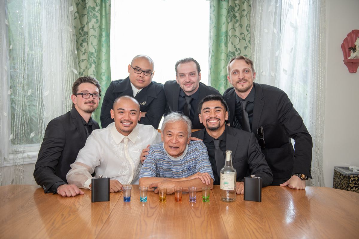 Seven men, including an elderly man at the center, gather around a table for a toast with shot glasses and bottles of liquor.