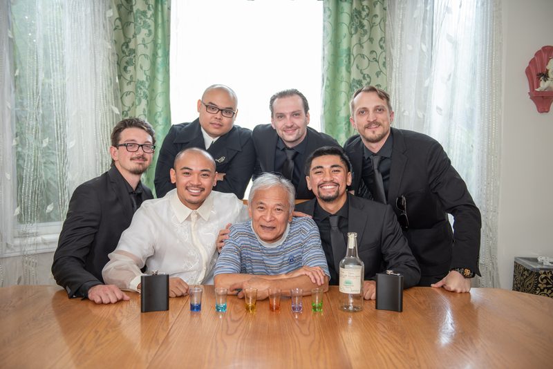 Seven men, including an elderly man at the center, gather around a table for a toast with shot glasses and bottles of liquor.
