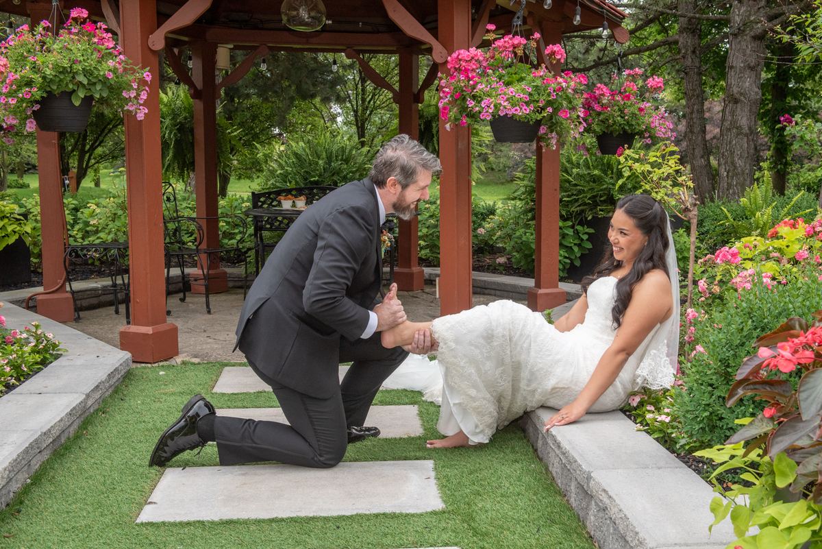 A groom in a dark suit kneels beside a bride in a white gown in a garden gazebo filled with pink flowers.