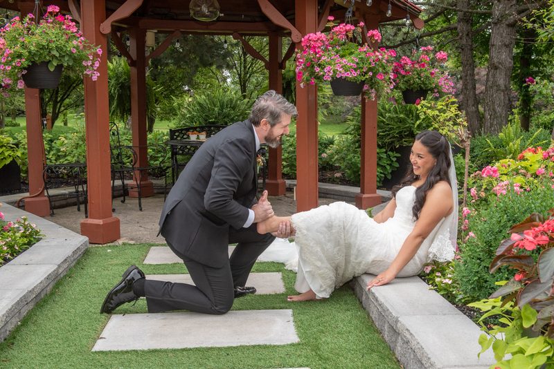 A groom in a dark suit kneels beside a bride in a white gown in a garden gazebo filled with pink flowers.