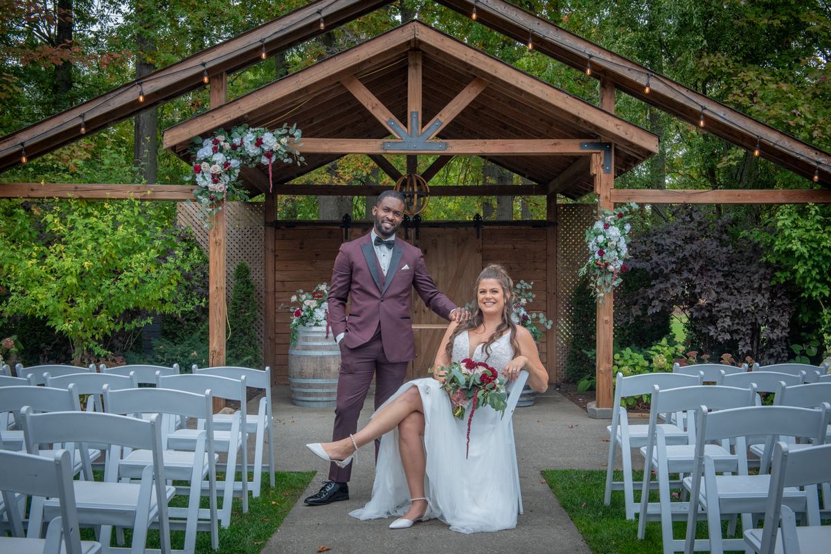 A smiling groom stands beside his seated bride under a floral-adorned wooden arbor in natural daylight.