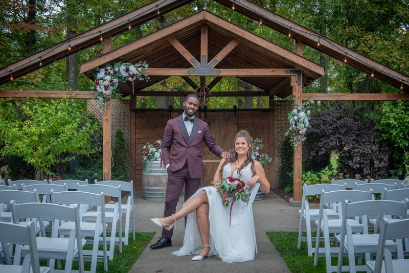 A smiling groom stands beside his seated bride under a floral-adorned wooden arbor in natural daylight.