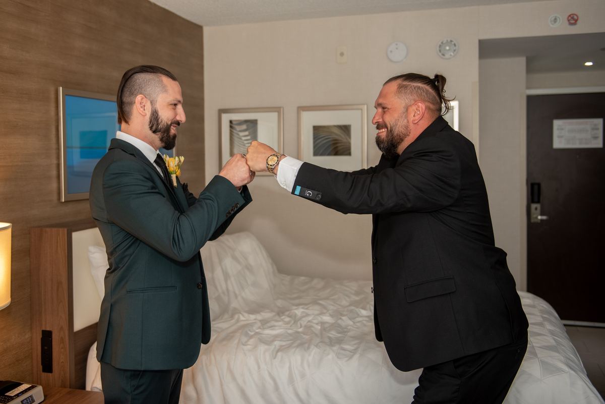 Two men in suits fist bump joyfully in a hotel room with warm lighting and one wearing a boutonnière.