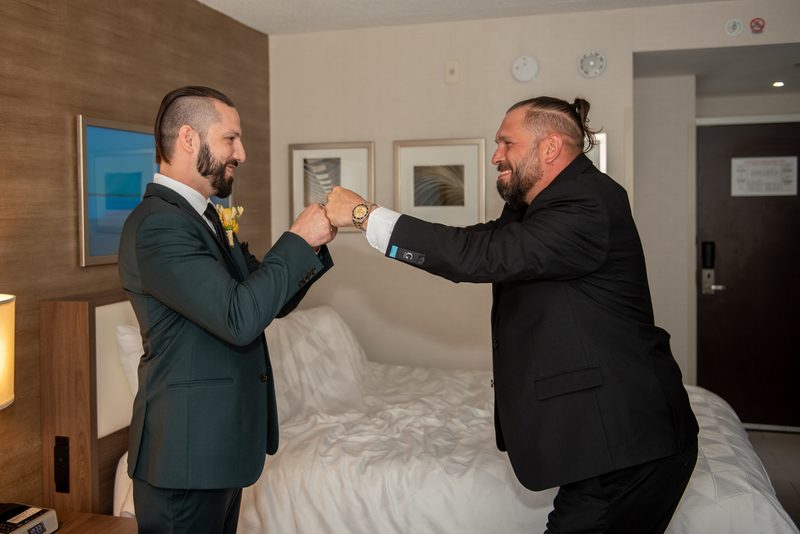 Two men in suits fist bump joyfully in a hotel room with warm lighting and one wearing a boutonnière.